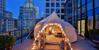 Tent with a bedroom set up on a downtown Chicago balcony with a cityscape in the background at nighttime.