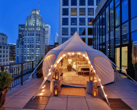 Tent with a bedroom set up on a downtown Chicago balcony with a cityscape in the background at nighttime