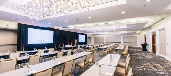 A large conference room with rows of tables, chairs and a large projector screen.