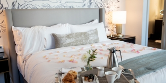 A tray of croissants and coffee laid on a hotel bed with pink rose petals spread over the duvet.