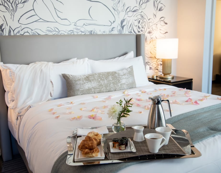 A tray of croissants and coffee laid on a hotel bed with pink rose petals spread over the duvet.